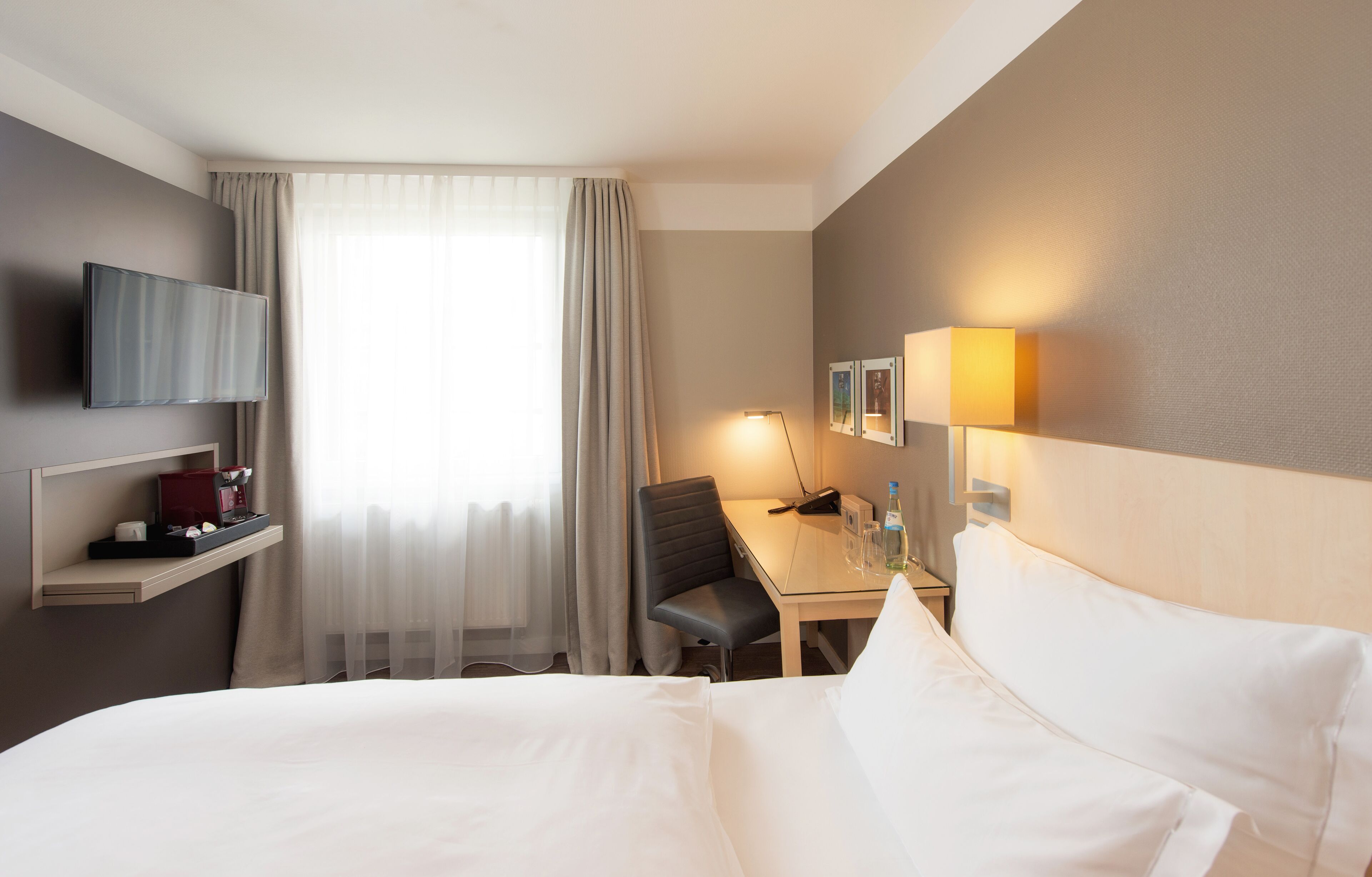 A hotel room with a bed, a desk, a chair, a TV, and a lamp.