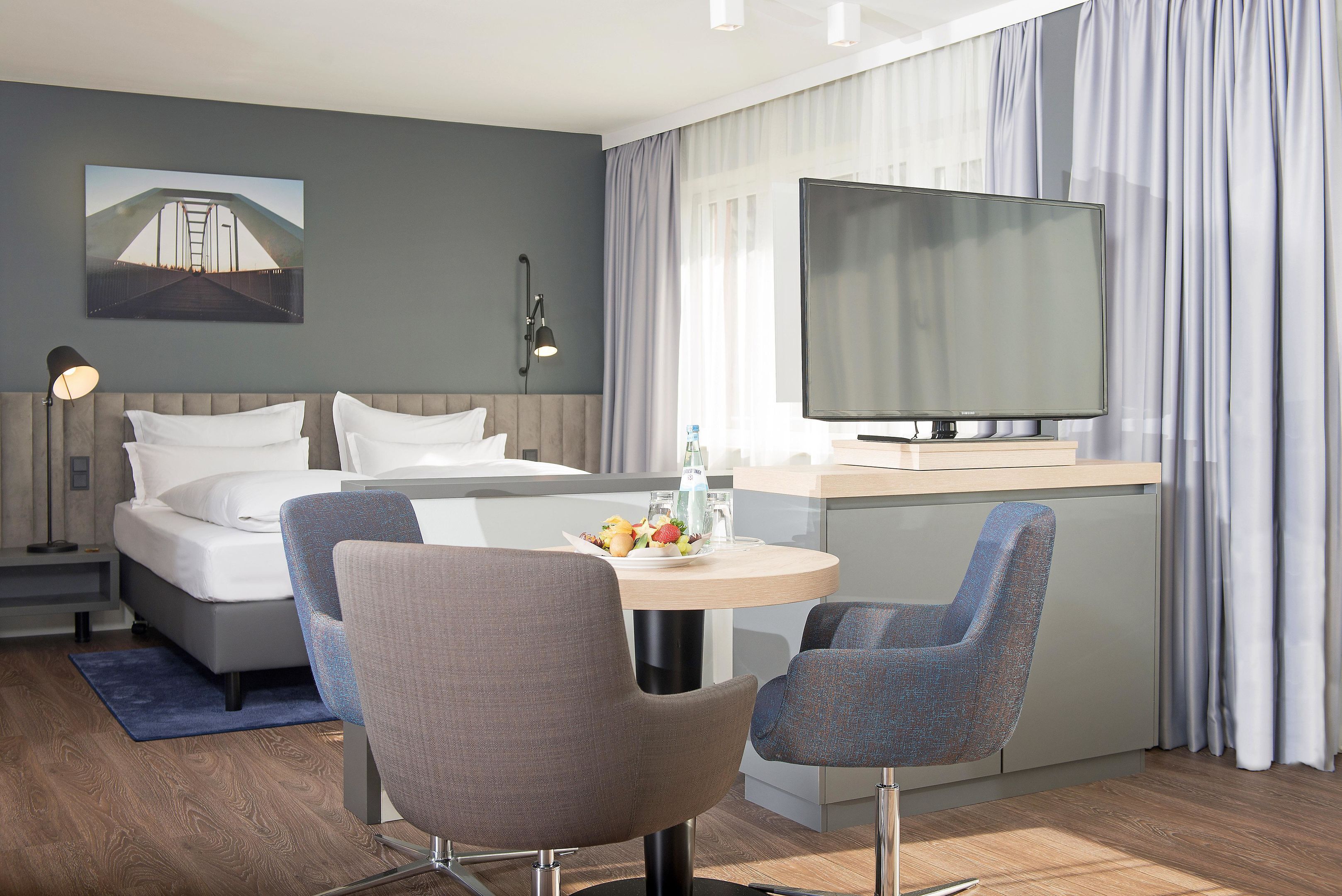 Una habitación de hotel moderna con una cama, un escritorio con televisión de pantalla plana, dos sillas, una mesa de comedor con una bandeja de fruta y un cuadro de un puente en la pared.