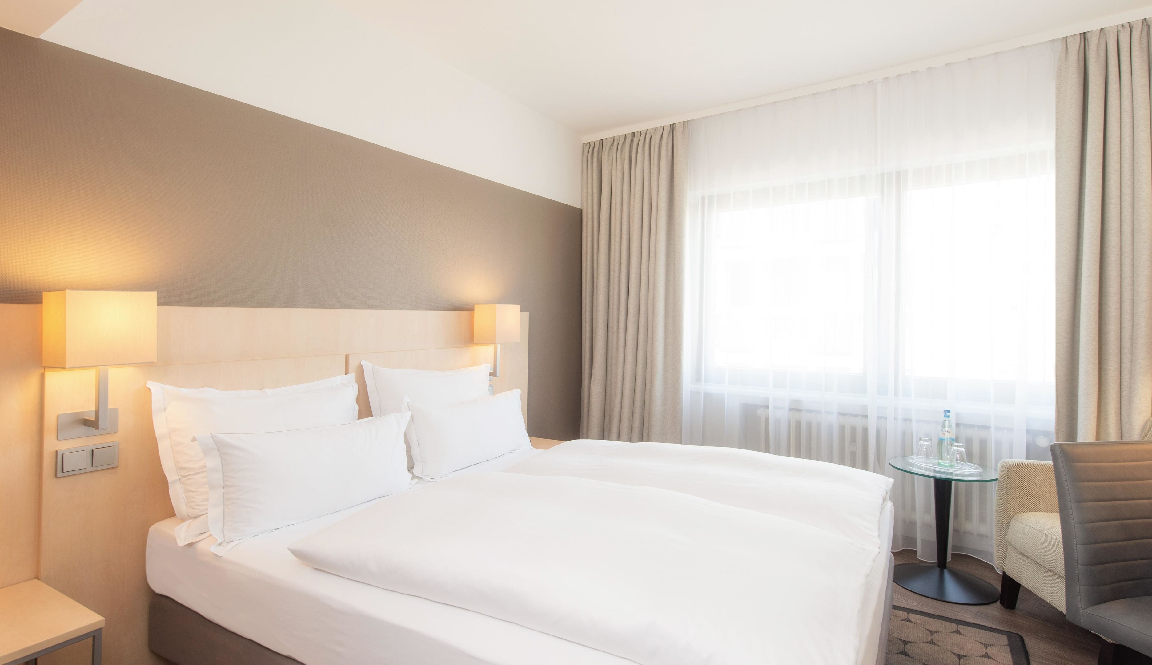 A hotel room with a large bed, two bedside lamps, a small table, a chair, and a large window with curtains.