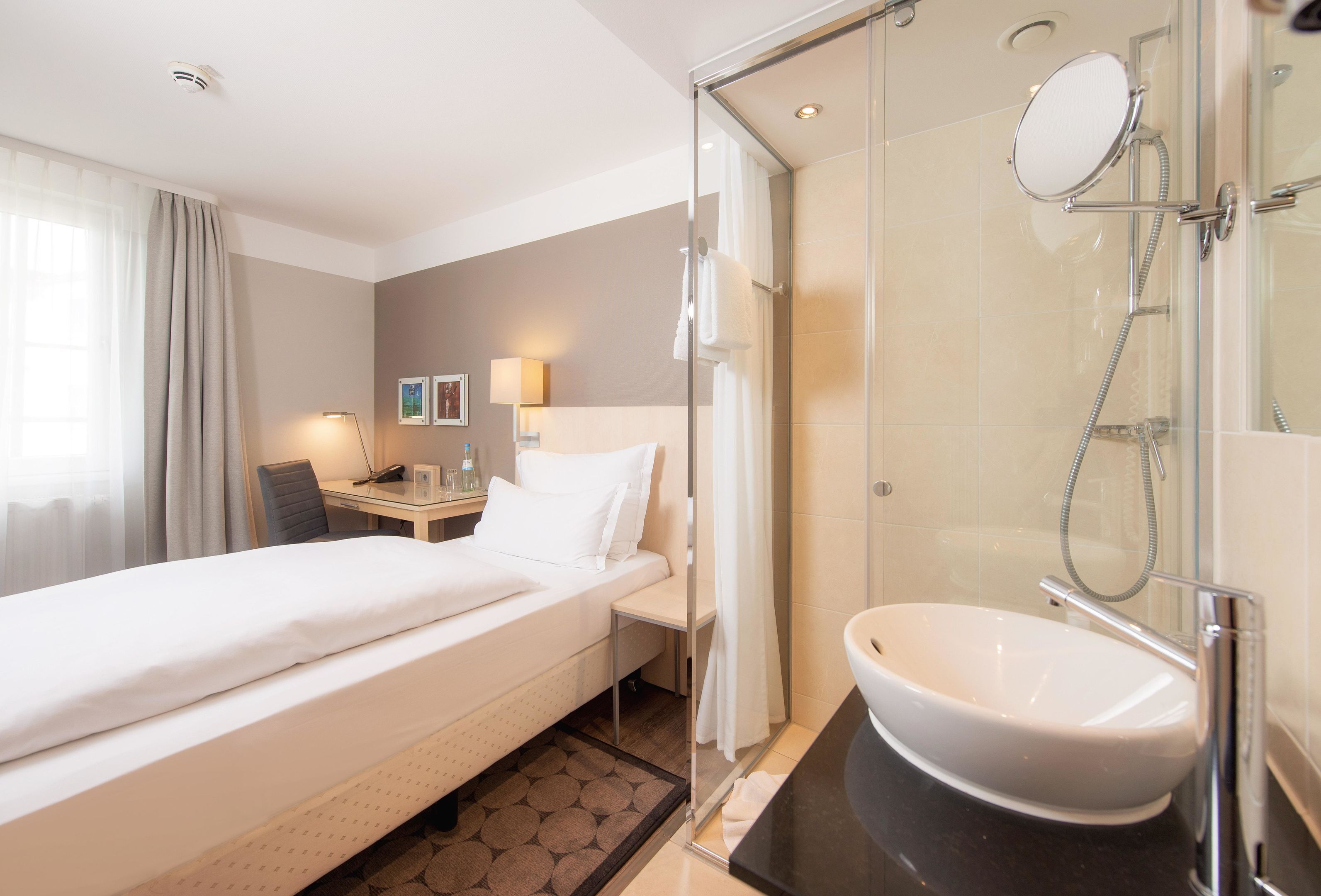 Ein Hotelzimmer mit Bett, Schreibtisch, Dusche und einem Badezimmer mit Waschbecken und Spiegel.