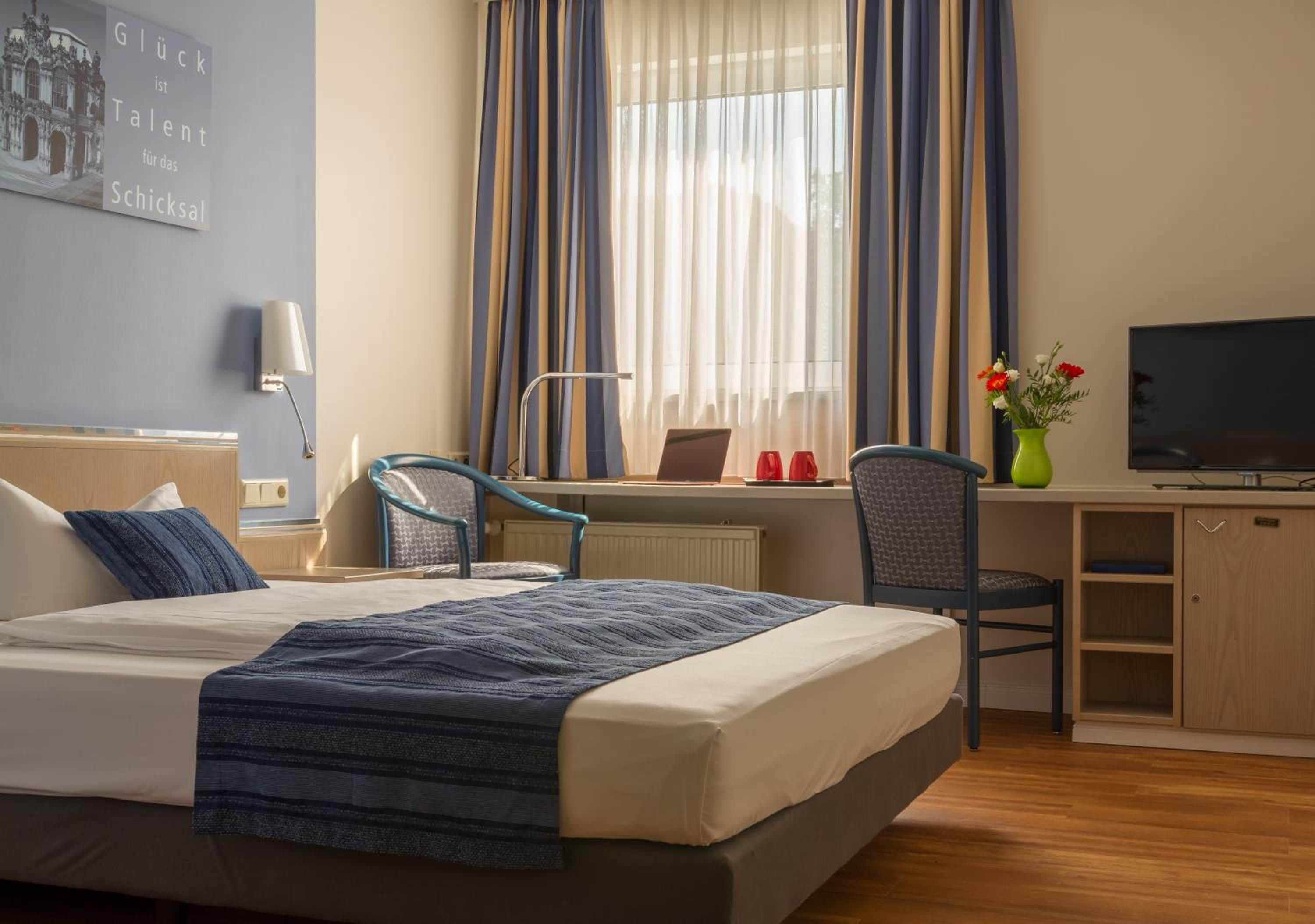 Habitación de hotel con cama, escritorio, silla, televisor y ventana con cortinas.