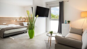 Habitación de hotel moderna con una cama, una planta verde, un sofá, un televisor y una ventana con cortinas.