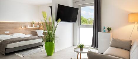 Habitación de hotel moderna con una cama, una planta verde, un sofá, un televisor y una ventana con cortinas.