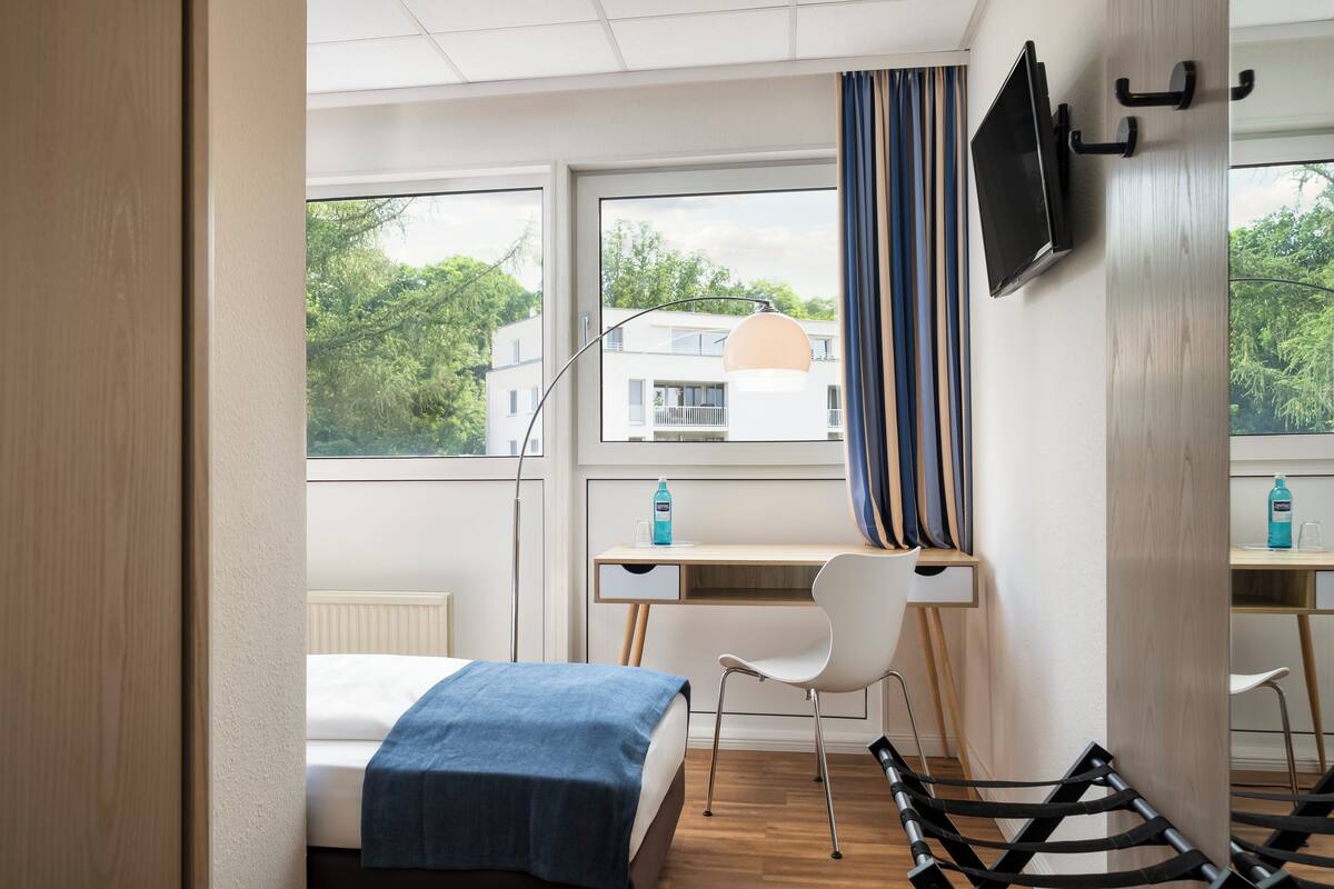A modern hotel room with a bed, a desk, a chair, a TV, and a window with curtains.