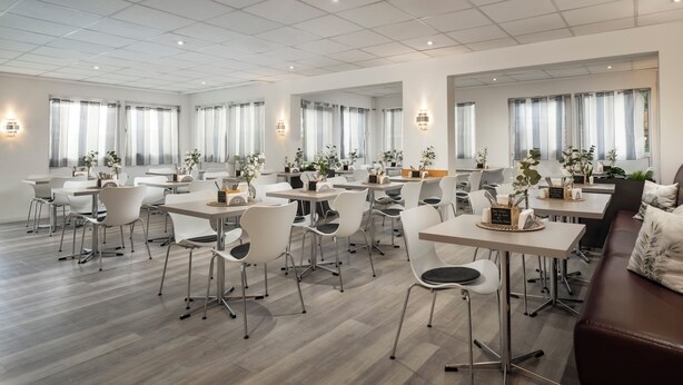 Un comedor moderno de hotel con varias mesas y sillas, ventanas amplias con cortinas y plantas en macetas.