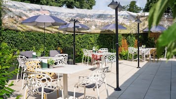 Een buitenpatio met witte metalen stoelen en tafels, parasols en een uitzicht op een stadslandschap door een decoratief scherm.