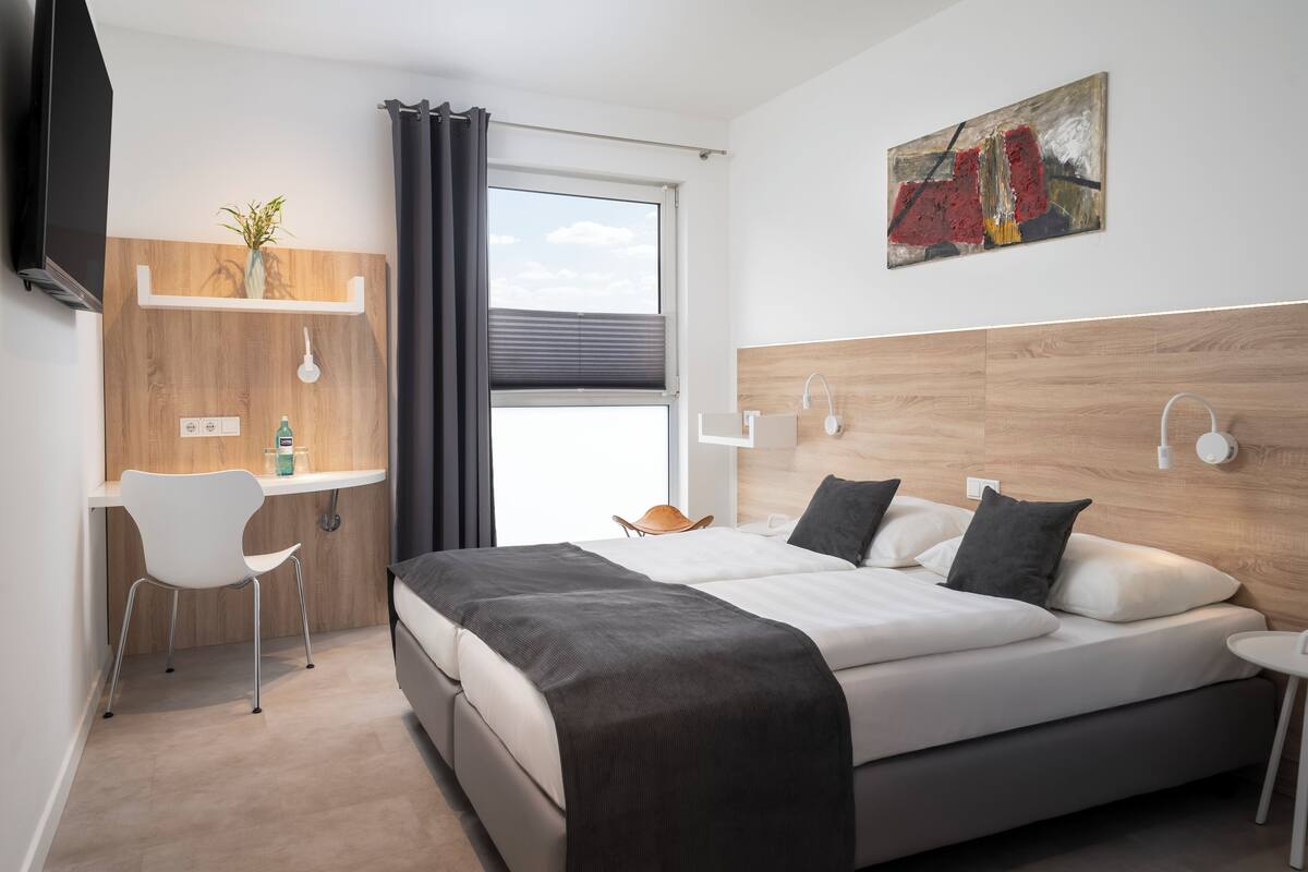 A modern hotel room with a bed, a desk, a chair, a TV, a window with curtains, and a painting on the wall.