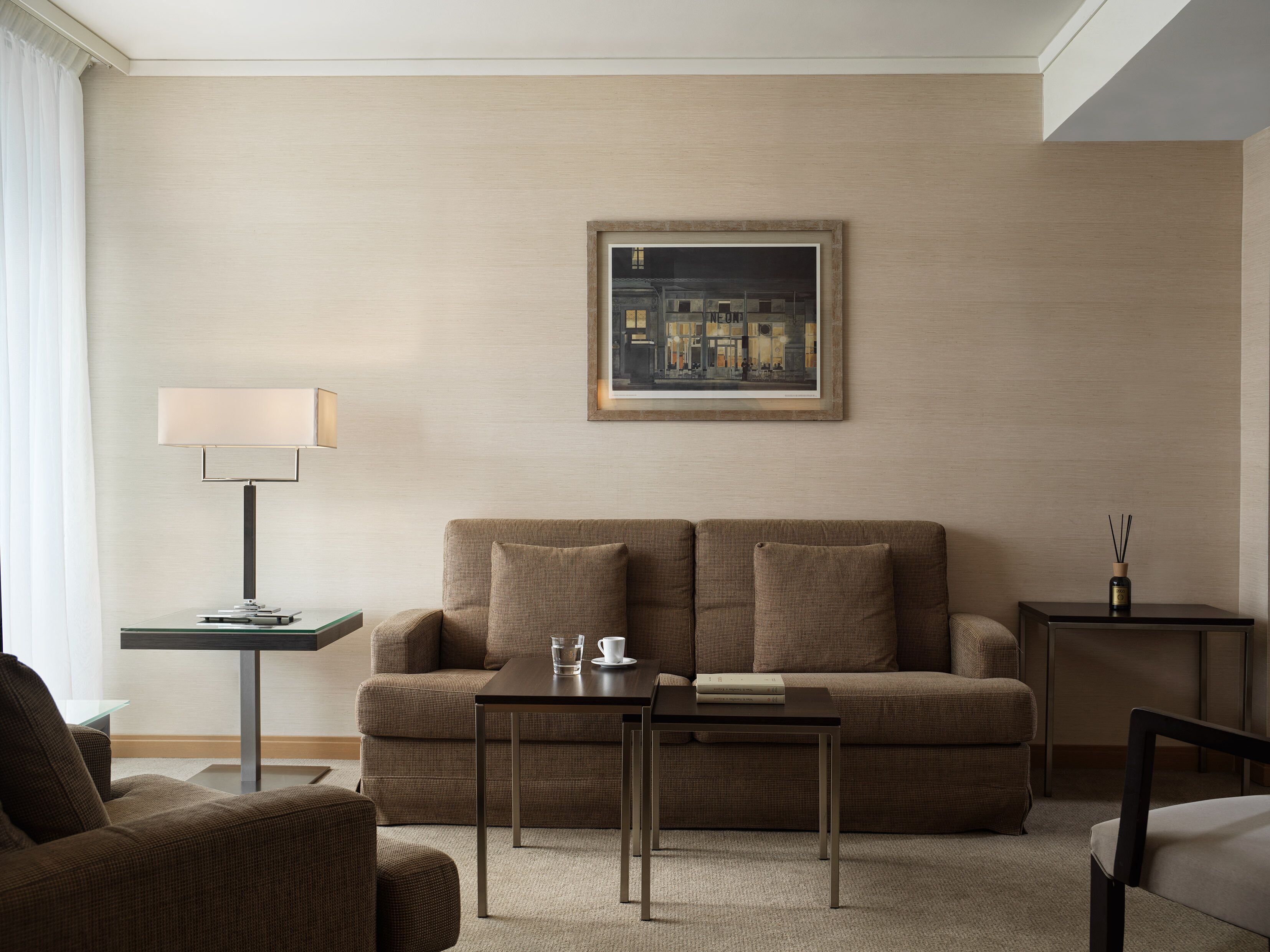 Una habitación de hotel moderna con un sofá beige, una mesita, una lámpara y un cuadro enmarcado en la pared.