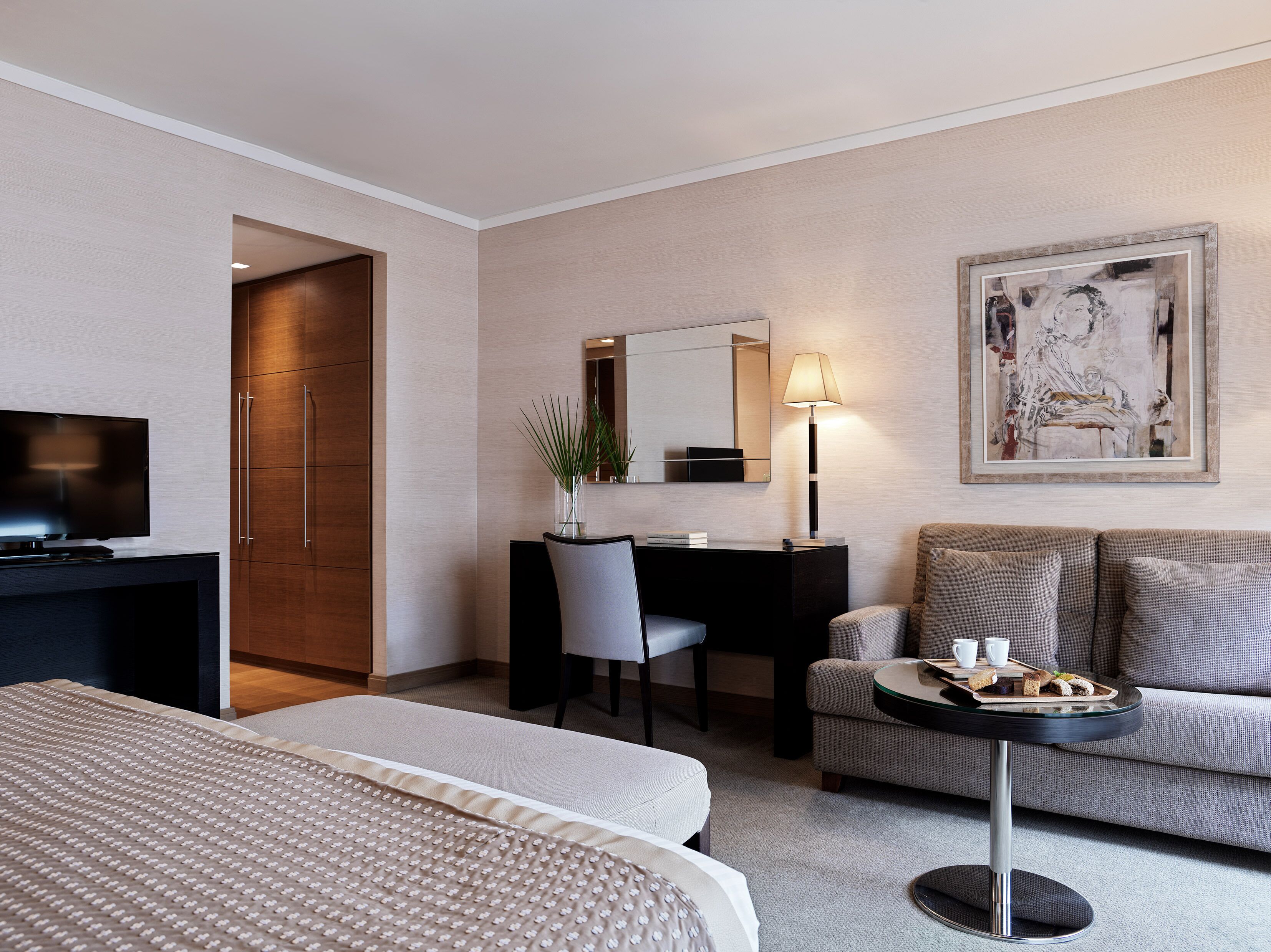 A hotel room with a bed, a desk with a chair, a sofa, a small table with a tray of food, a lamp, and a painting on the wall.