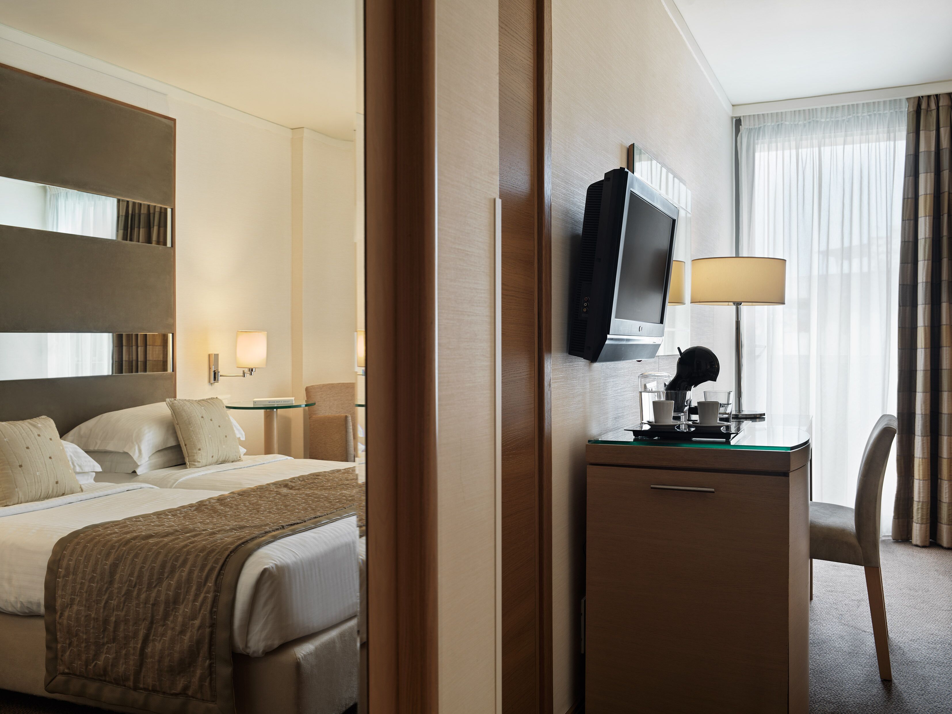 A hotel room with a bed, a desk with a TV, a lamp, and a chair.