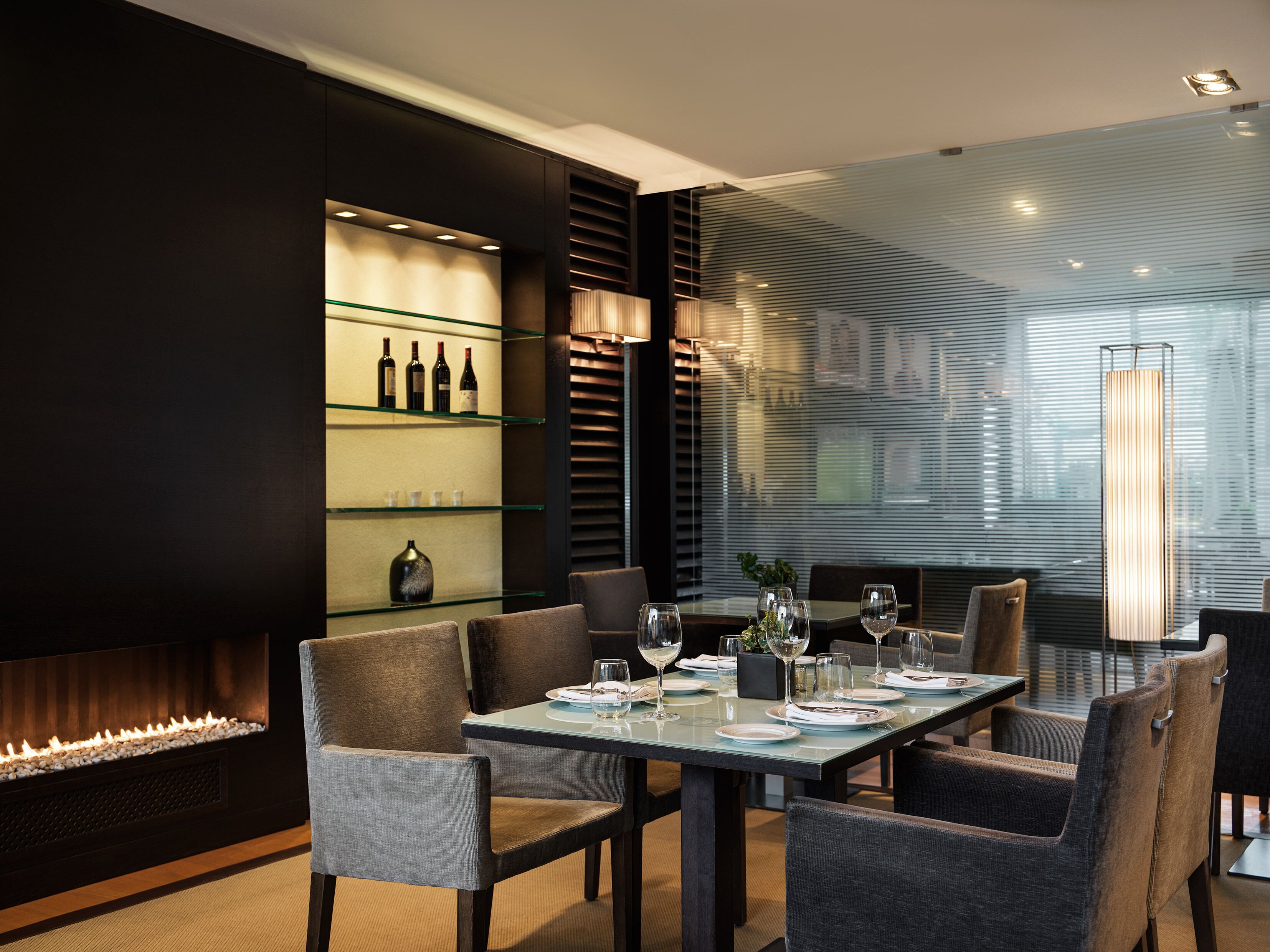 Un comedor moderno con una mesa de vidrio, sillas tapizadas, un espacio para exhibir vinos y una chimenea.