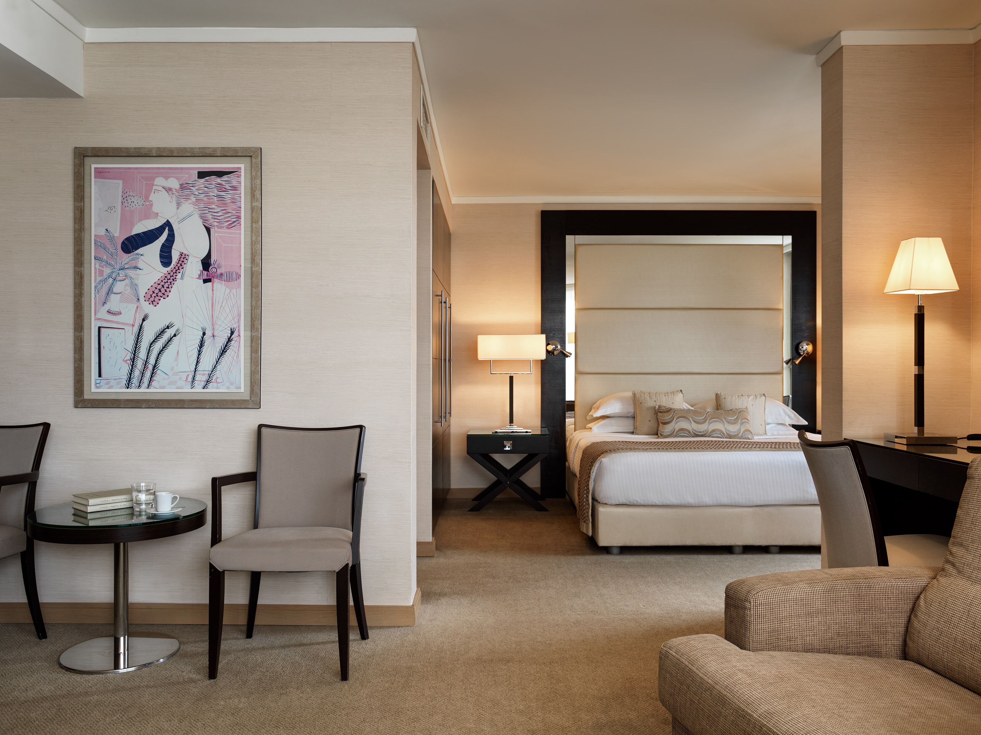 A hotel room with a bed, a desk, a chair, a small table, and a painting on the wall.
