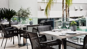An outdoor dining area with wicker chairs and tables, potted plants, and a modern building in the background. - Galaxy Hotel Iraklio (Heraklion)