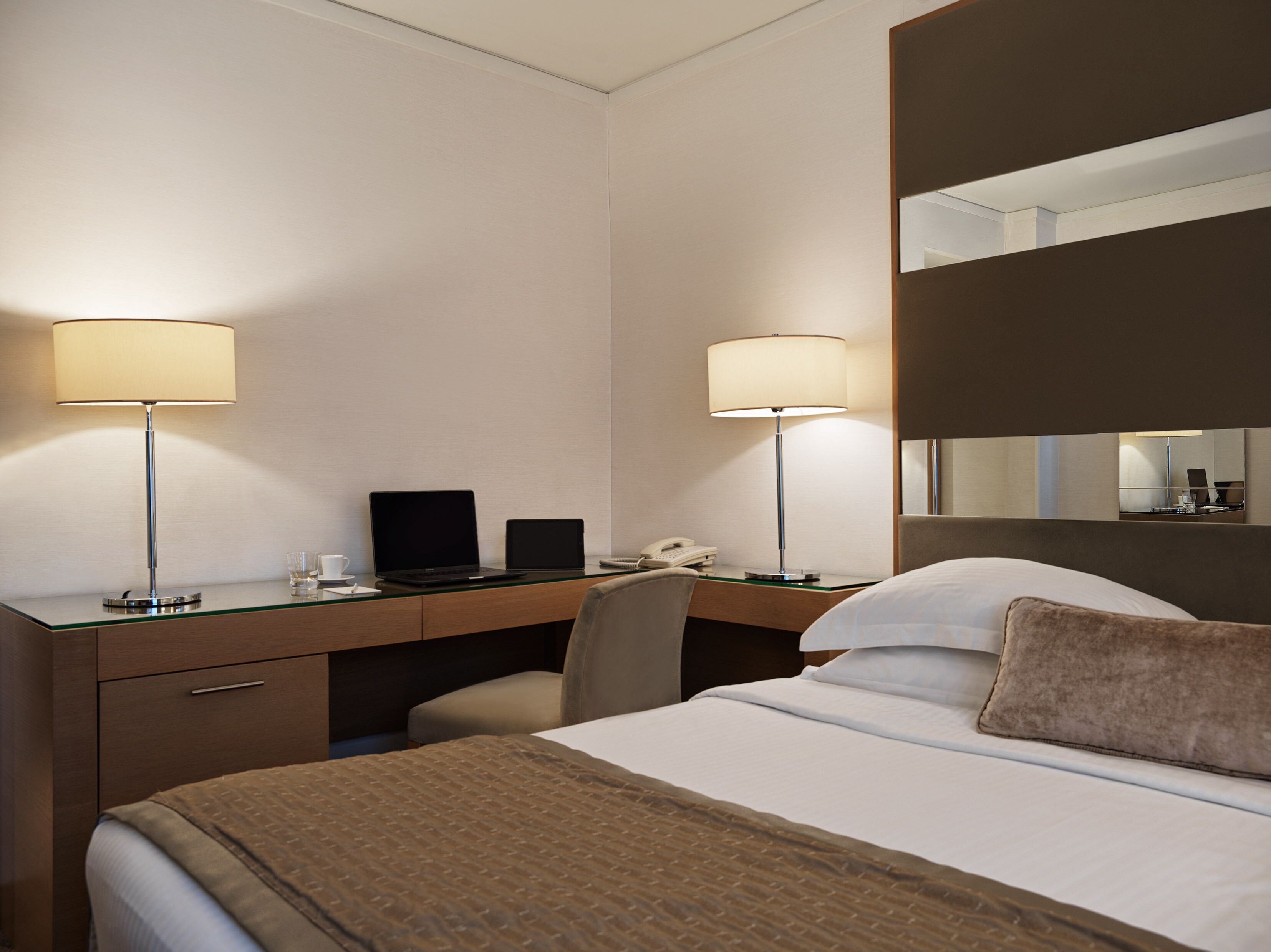 A hotel room with a bed, a desk with two laptops, a telephone, and two lamps.