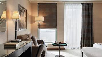 Una habitación de hotel moderna con un sofá, una pequeña mesa redonda con una bandeja, un escritorio con una lámpara y un amplio ventanal con cortinas.