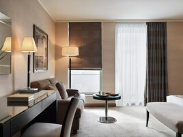 Una habitación de hotel moderna con un sofá, una pequeña mesa redonda con una bandeja, un escritorio con una lámpara y un amplio ventanal con cortinas.