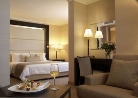 Quarto de hotel com cama, mesa de cabeceira com abajur, uma cadeira, mesa de jantar com prato de comida e taça de vinho, e um espelho na parede.
