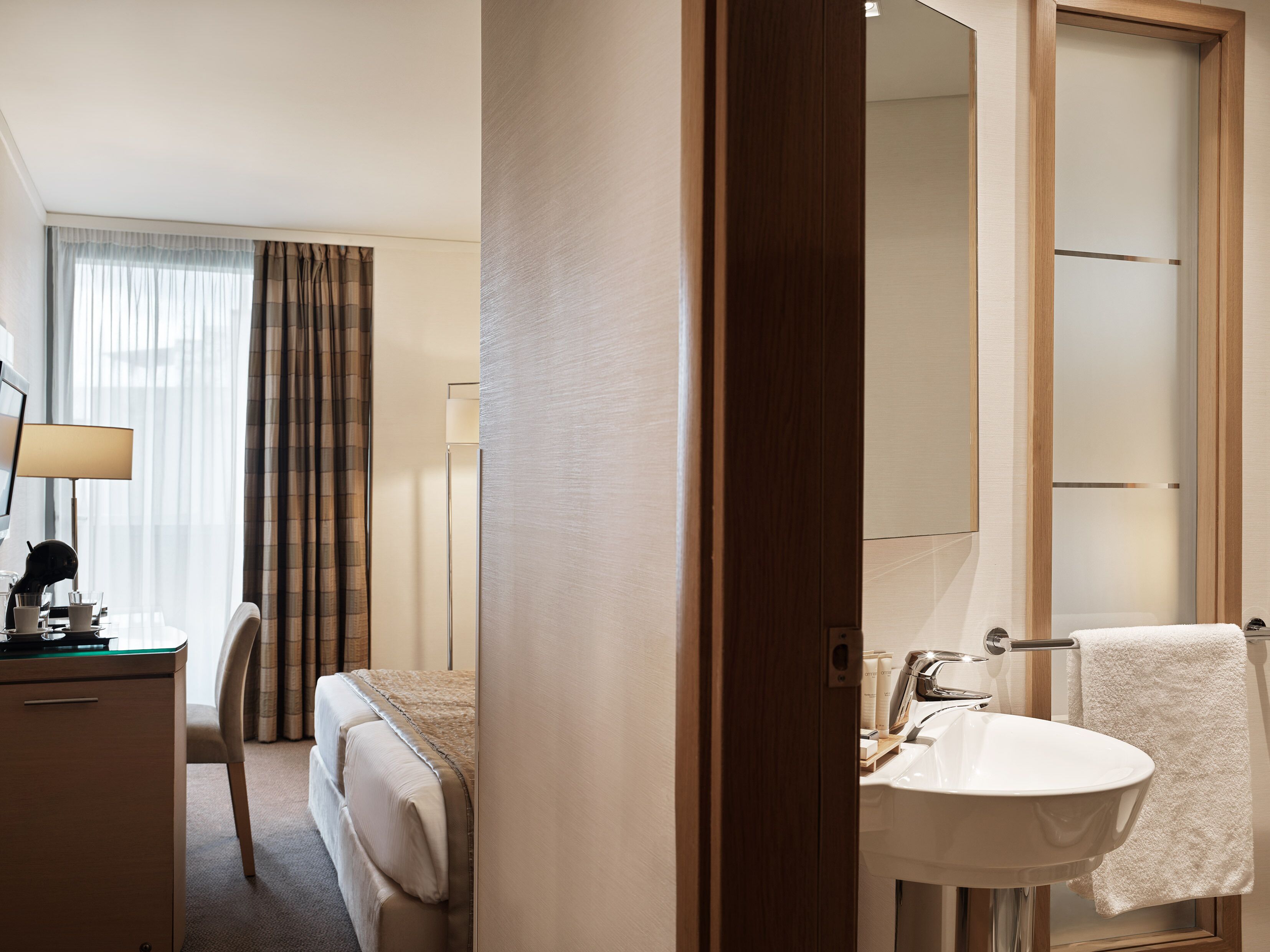 Habitación de hotel con cama, escritorio con lámpara, mesita de noche, espejo y baño con lavamanos y toalla.