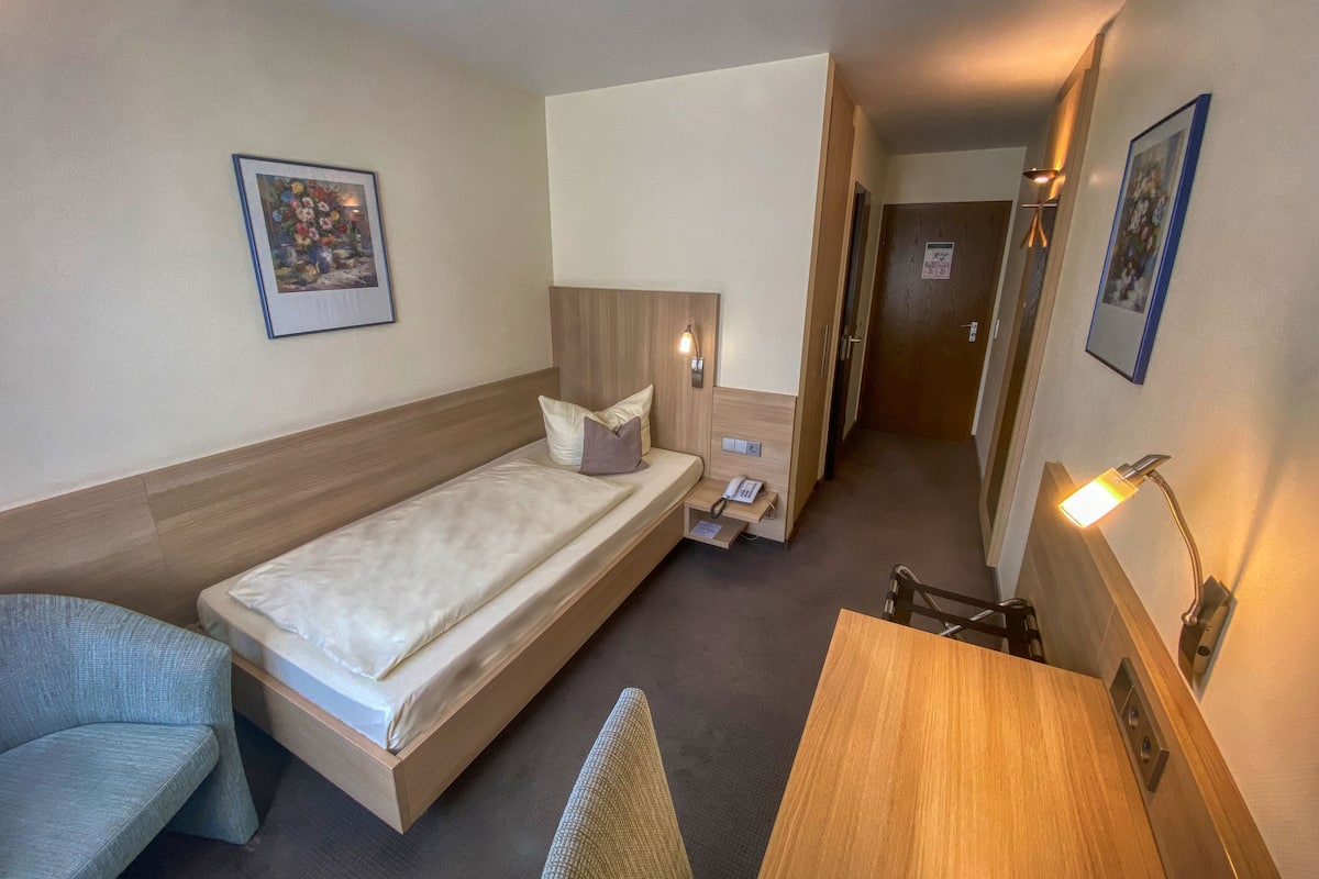 A compact hotel room with a single bed, a desk, a chair, a bedside table, a lamp, and a phone.