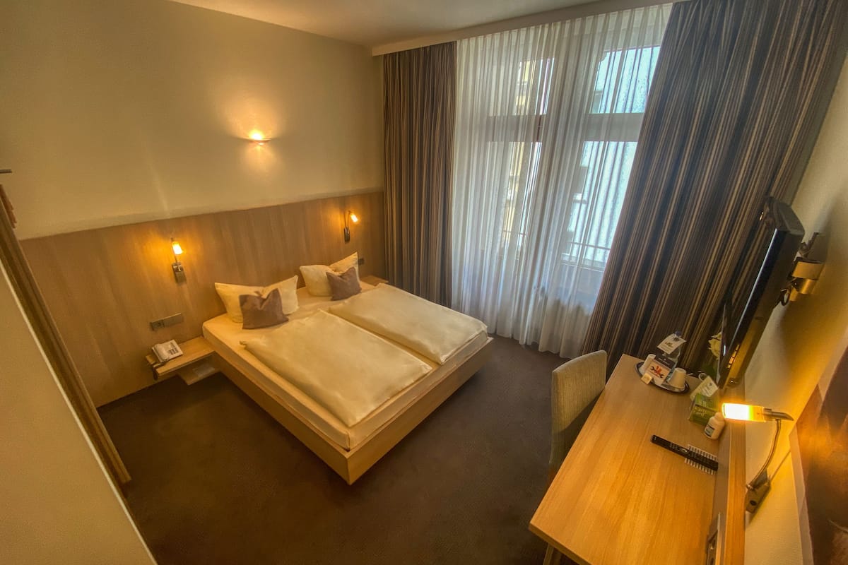A hotel room with a double bed, bedside tables, a desk with a chair, a coffee maker, and a window with curtains.
