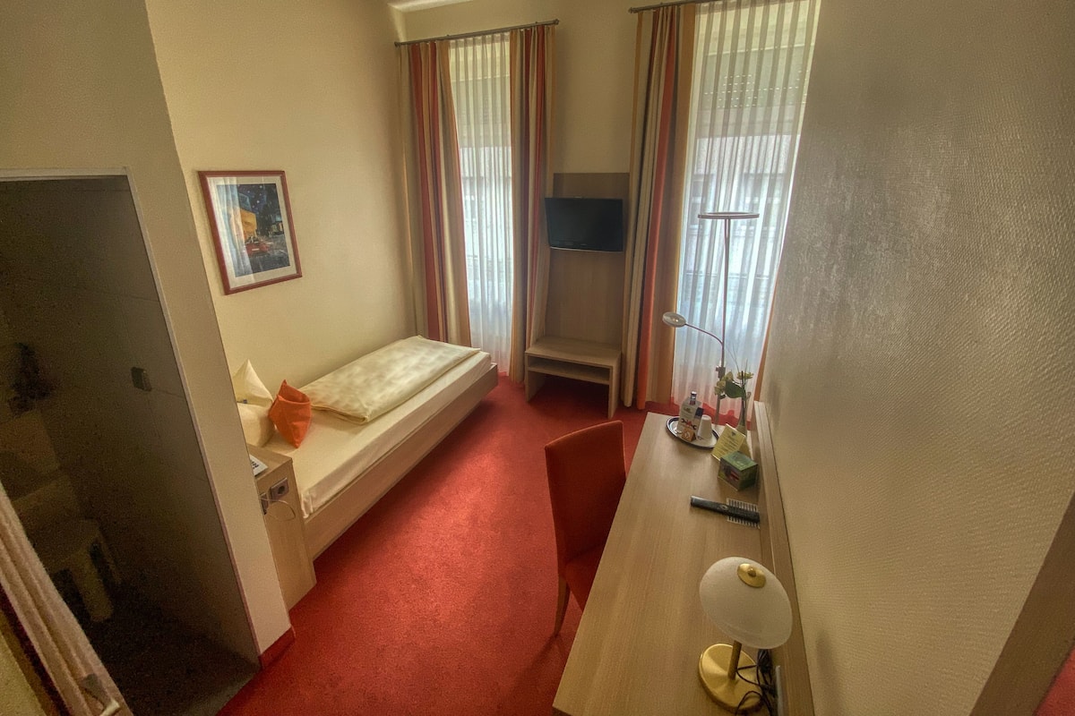 A small, single-bed room with a desk, a chair, a bedside table, a lamp, and a window with curtains.