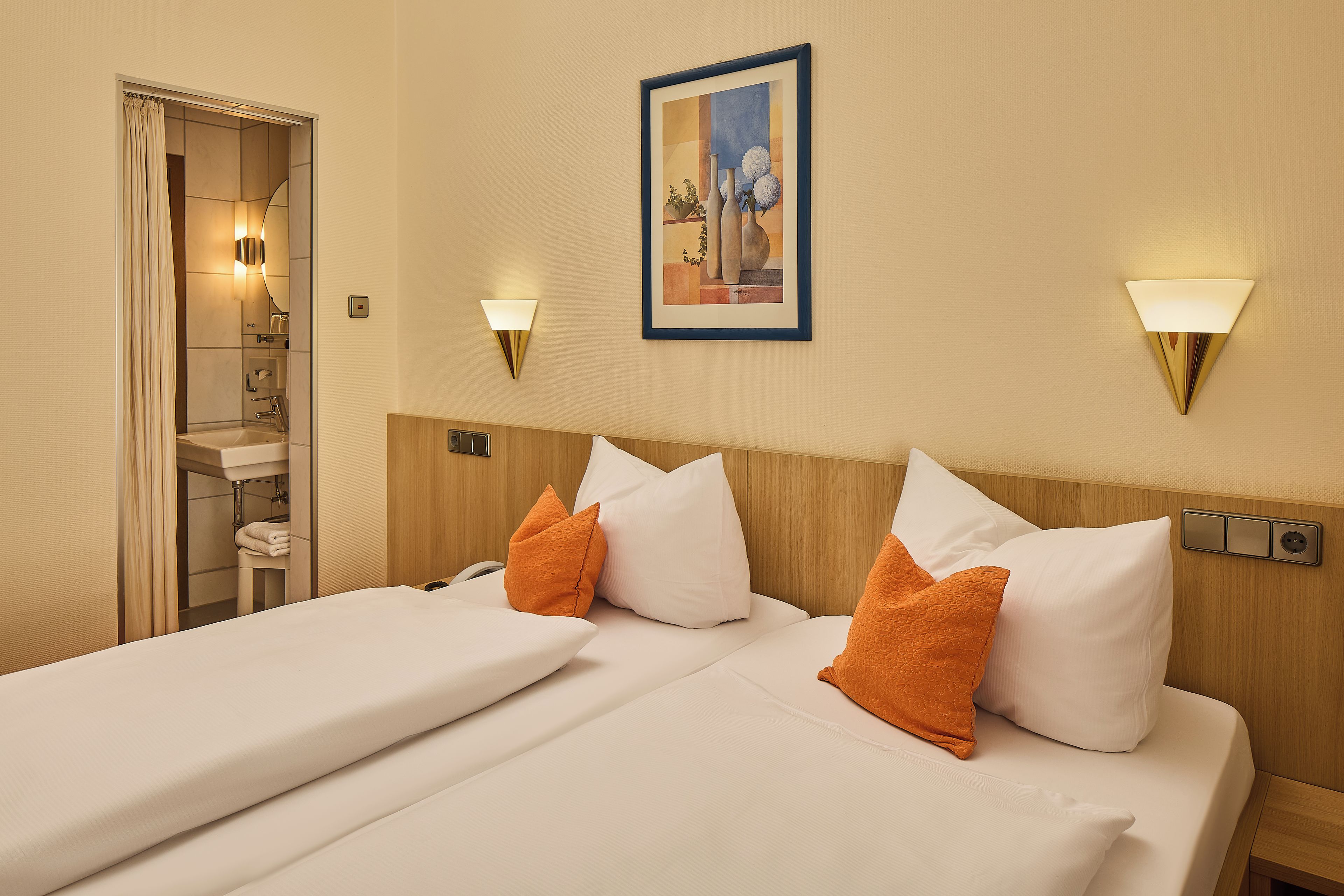 A hotel room with a bed, two pillows, a nightstand, a wall-mounted lamp, and a painting.