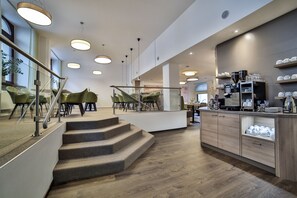 A modern interior with a staircase leading to a seating area, a kitchen counter with coffee-making equipment, and a view of the outdoors. - City Partner Hotel Berliner Hof (Karlsruhe)