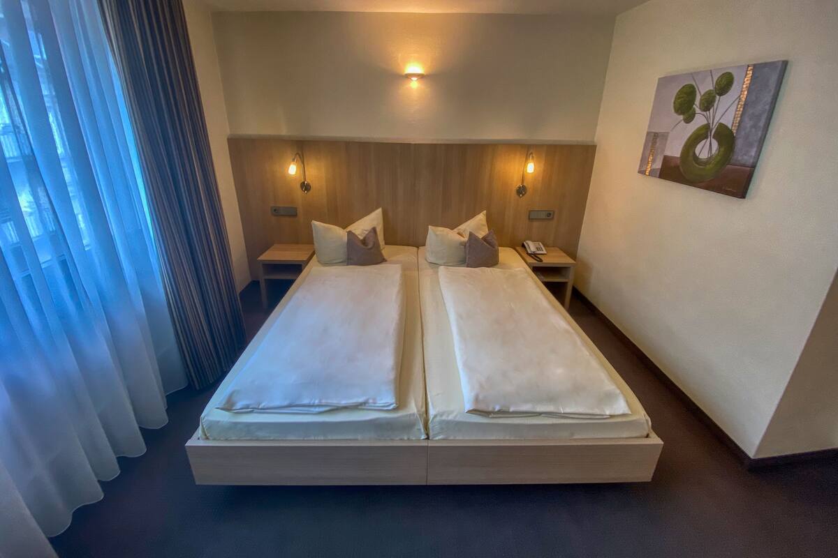 A double bed with white linens, two bedside tables with lamps, a wall-mounted painting, and a window with sheer curtains.