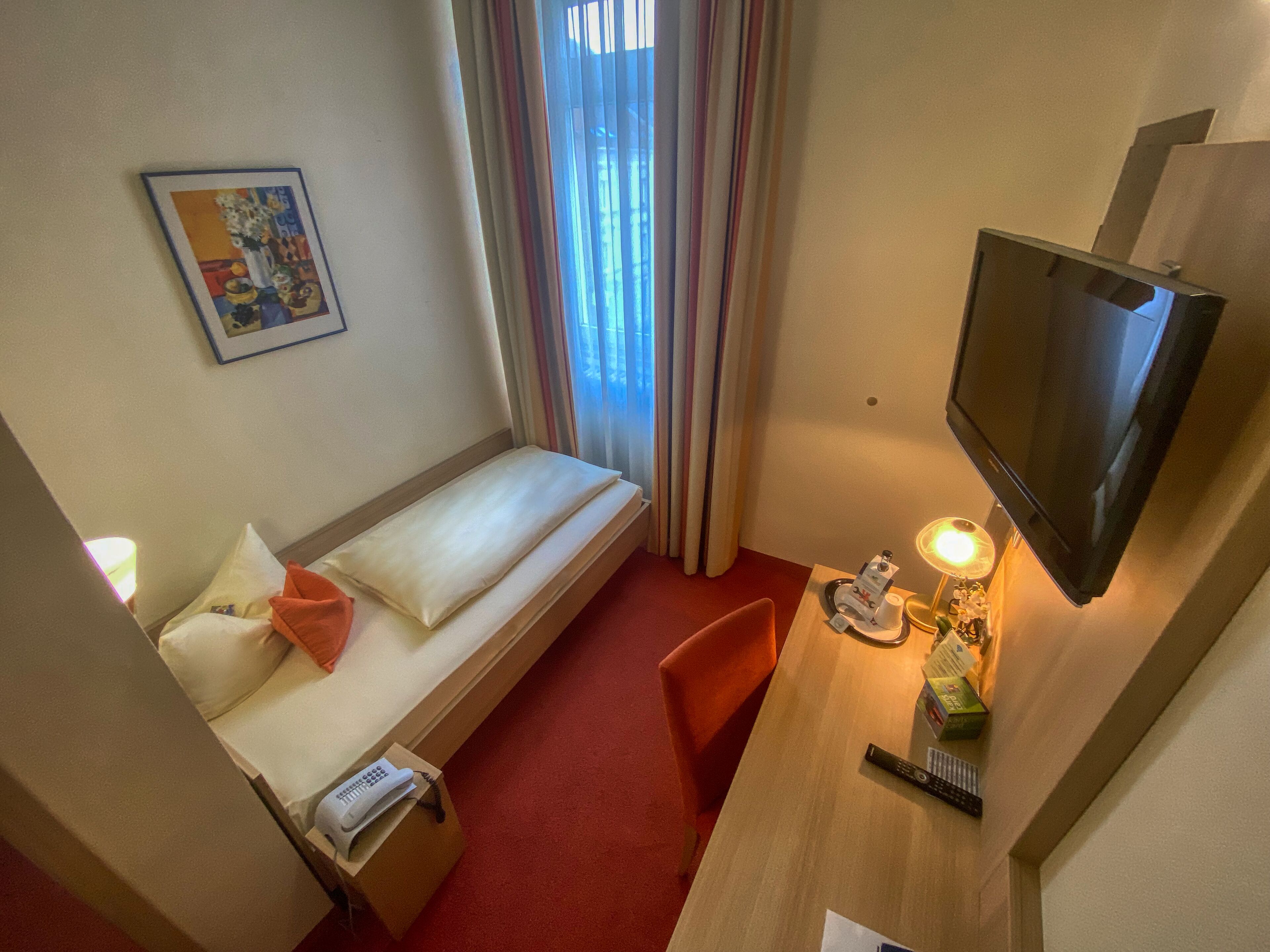 Quarto de hotel pequeno com uma cama de solteiro, uma escrivaninha com abajur, televisão e telefone.