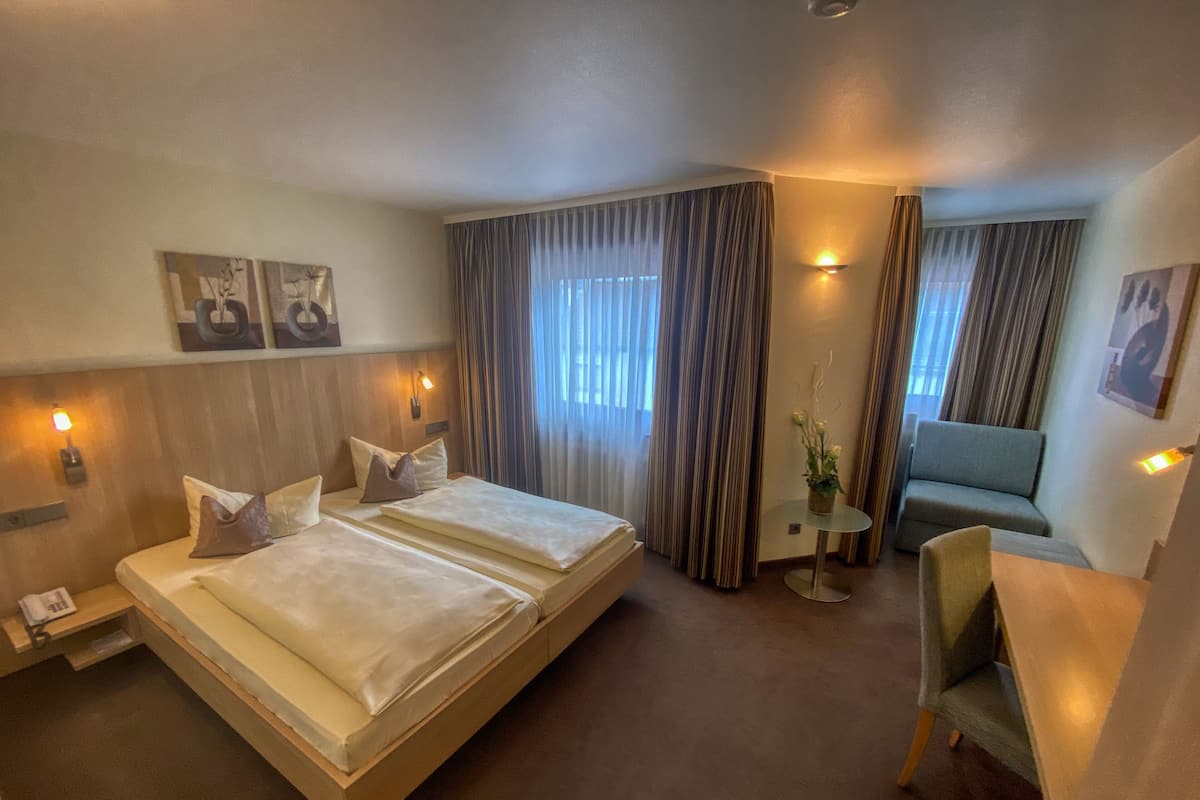 A hotel room with a large bed, a desk, and a chair. There are curtains, wall art, and a small table with a plant.