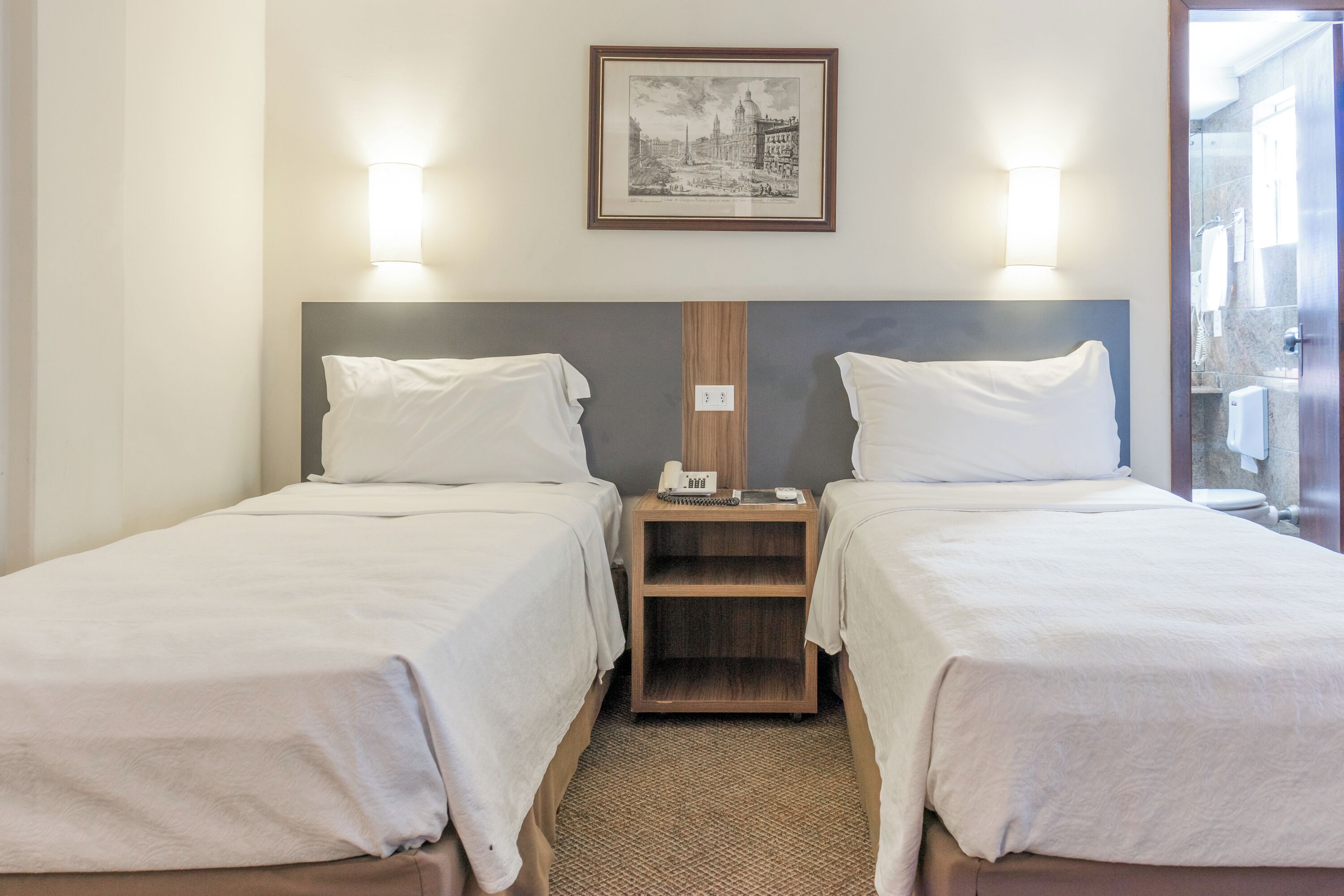 A double room with two beds, a wooden nightstand between them, a framed picture on the wall, and a bathroom visible through an open door.