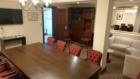 A conference room with a large wooden table, six red upholstered chairs, a television, and a bookshelf in the background. - Hotel Lancaster (Curitiba)