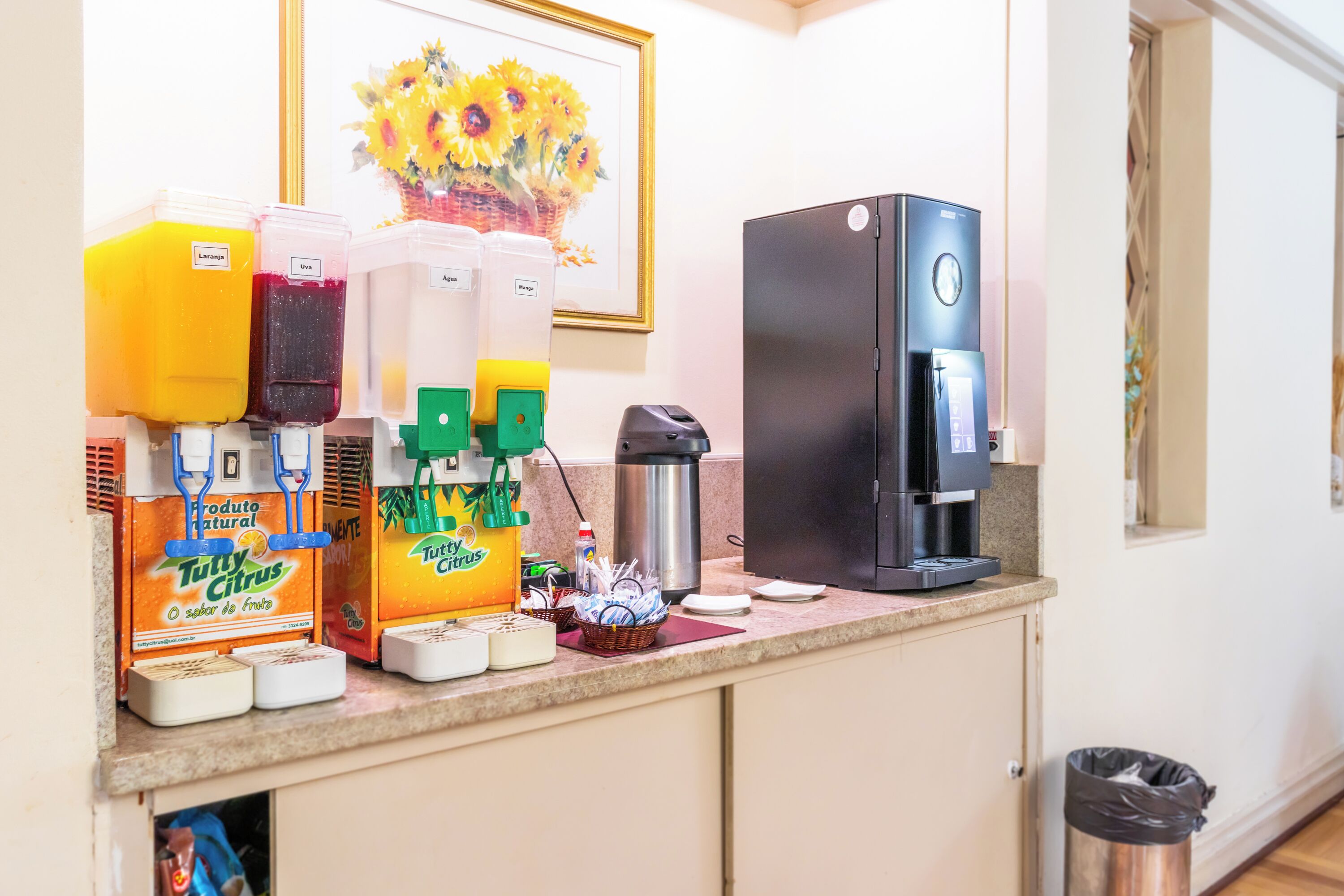 A kitchen counter with a coffee maker, a refrigerator, and juice dispensers.