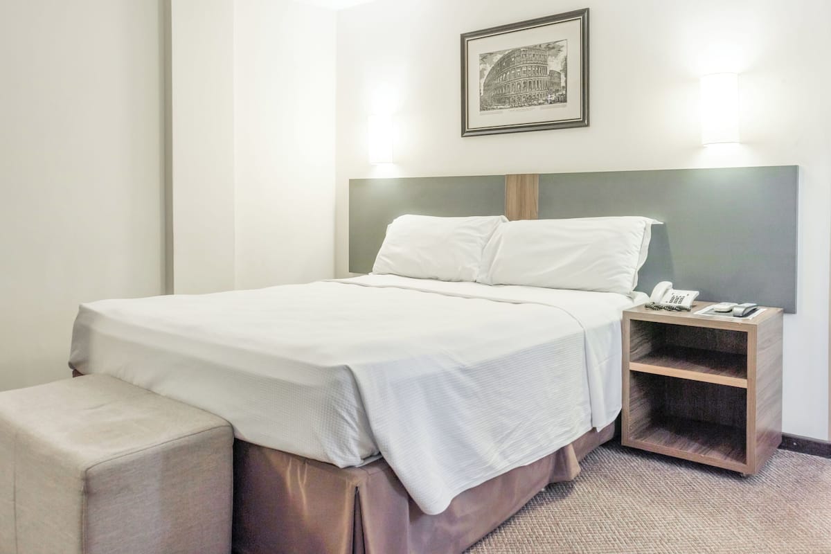 Habitación de hotel con una cama grande, una mesita de noche con lámpara, un pequeño banco y un cuadro enmarcado en la pared.
