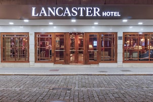 El exterior del Hotel Lancaster, con un letrero destacado, amplias ventanas de vidrio y una calle adoquinada al frente.