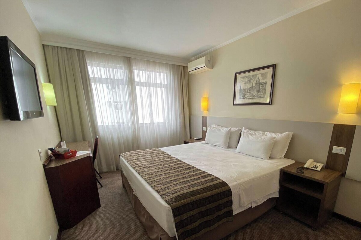 Une chambre d’hôtel avec un lit, un bureau, une chaise, une télévision et un tableau encadré au mur.