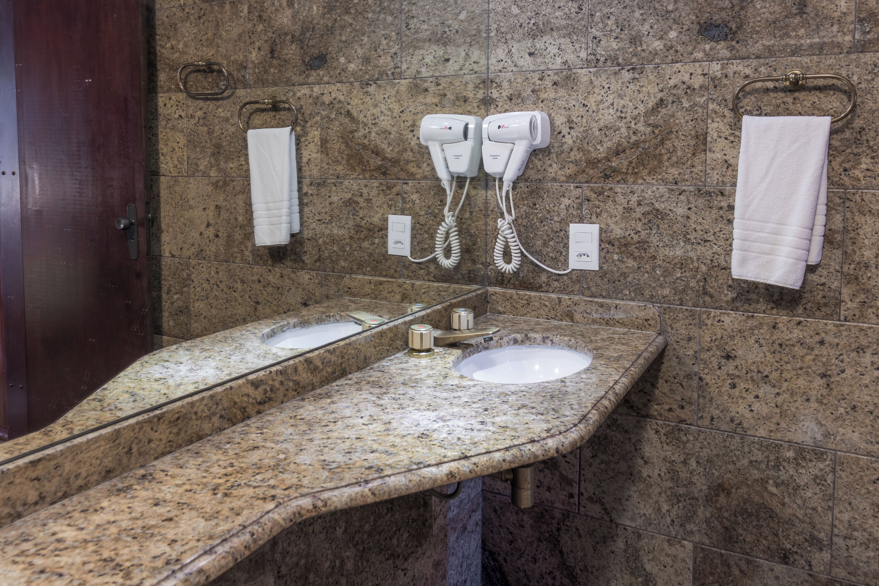 Casa de banho com duas pias, um espelho e dois secadores de cabelo fixados na parede.