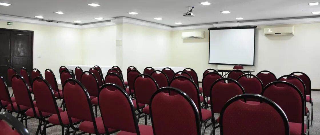 Un salón de conferencias con filas de sillas rojas, una pizarra y una pantalla de proyector.
