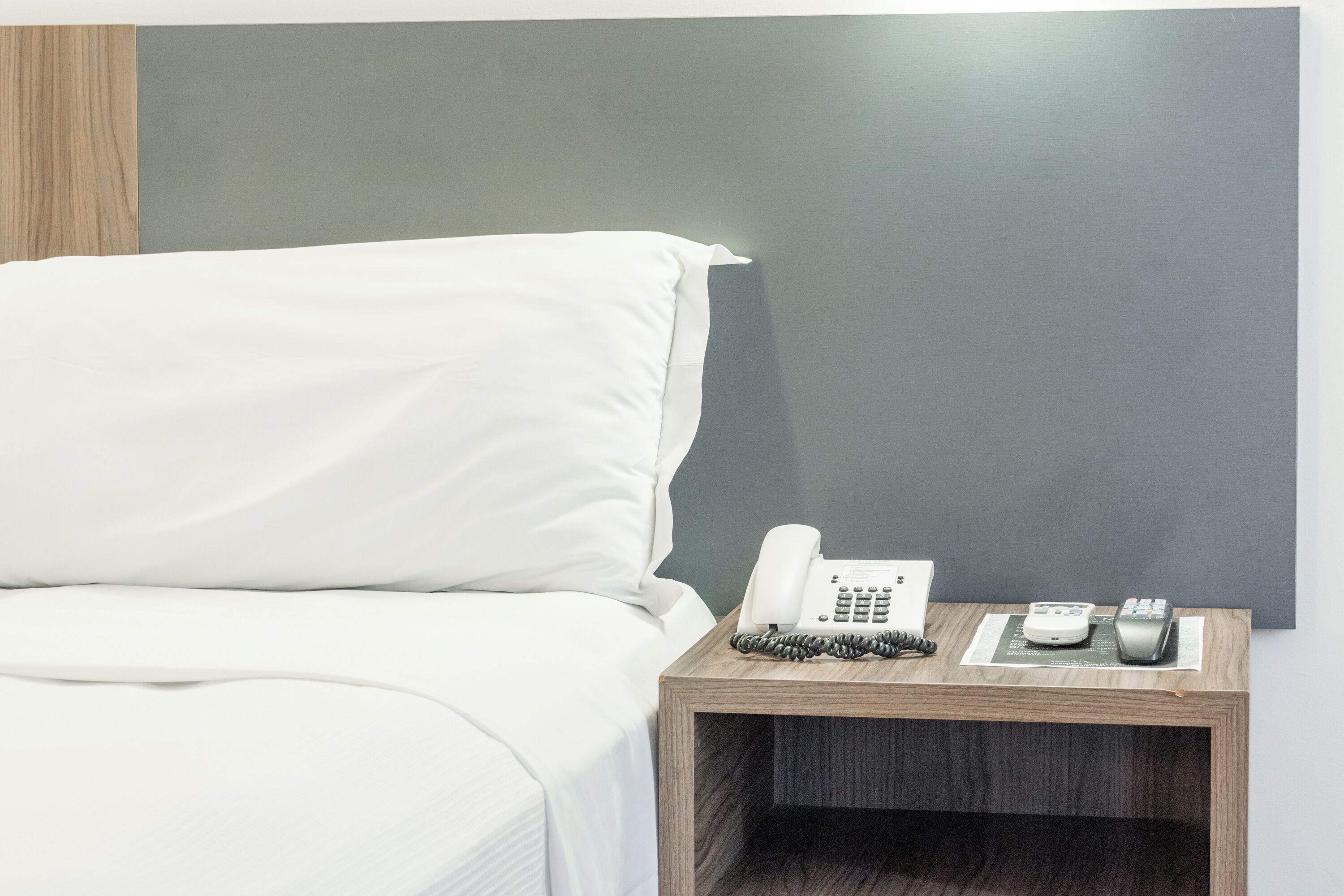 Un lit bien fait, avec du linge de lit blanc, une table de chevet en bois sur laquelle se trouvent un téléphone et une télécommande, et un mur gris foncé en arrière-plan.