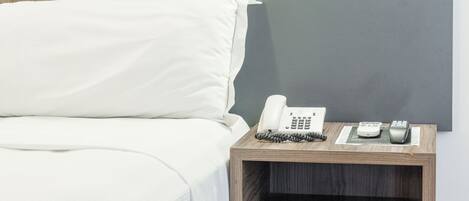 Una cama bien hecha con sábanas blancas, una mesita de noche de madera con un teléfono y un control remoto, y una pared gris oscuro al fondo.
