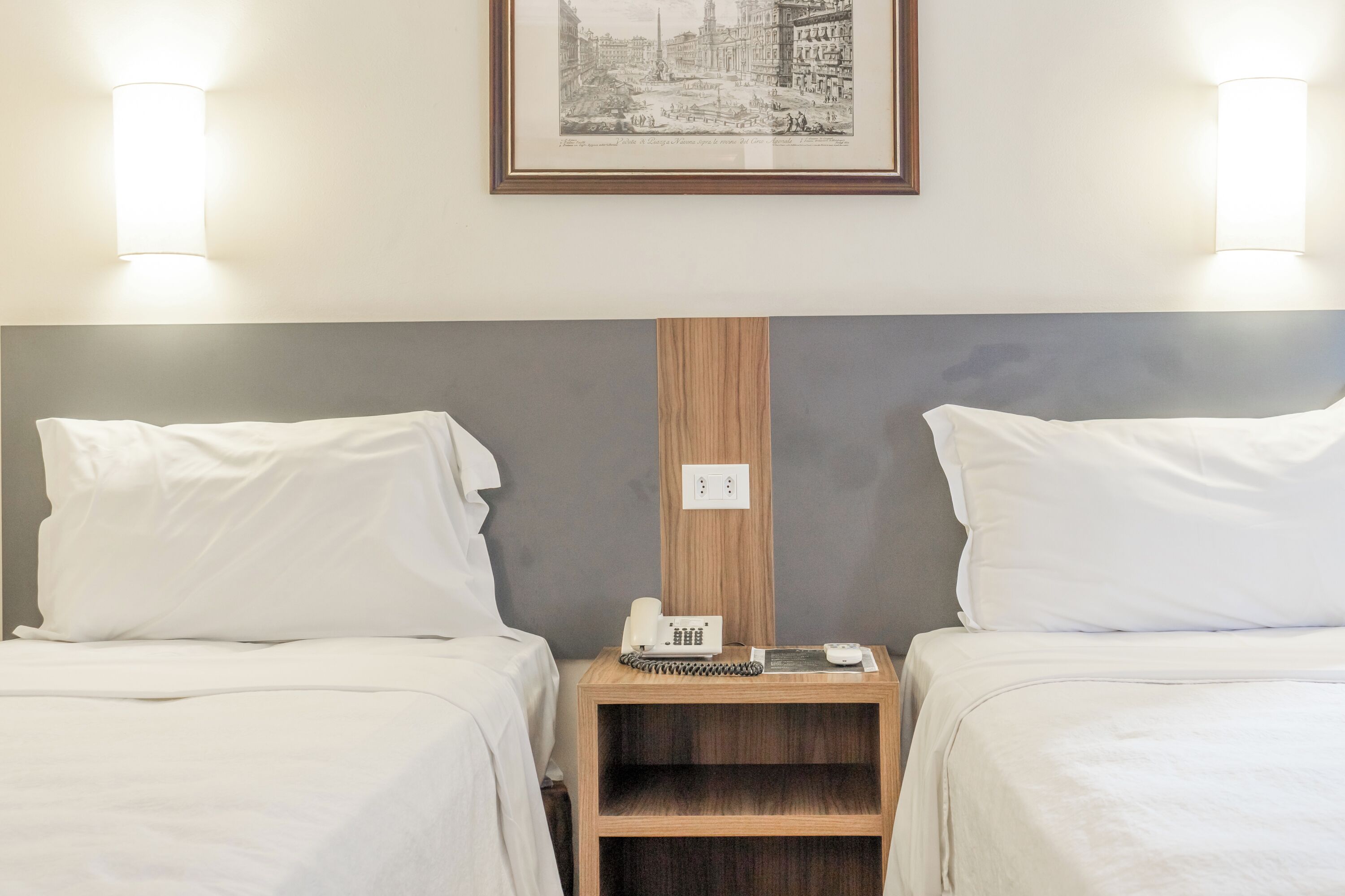 A hotel room with two beds, a wooden nightstand with a phone and a notepad, and a framed cityscape picture on the wall.