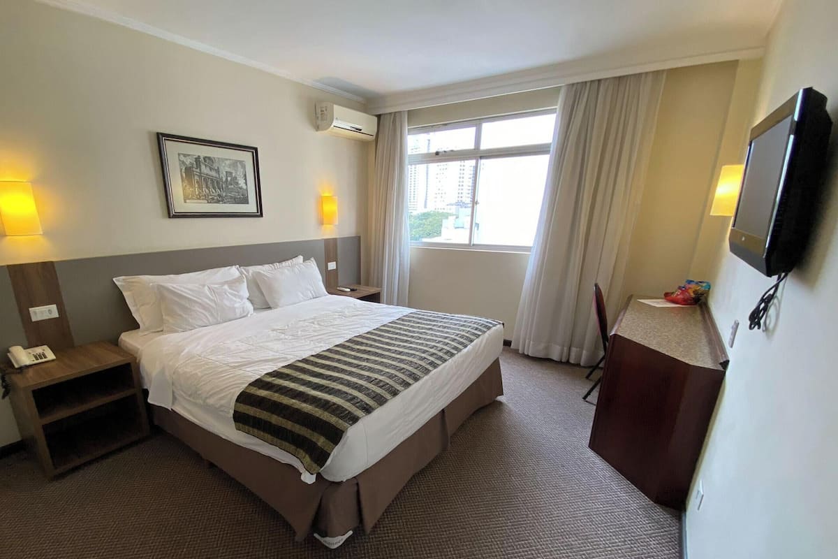 Habitación de hotel con cama, mesita de noche, lámpara, aire acondicionado, televisor y ventana con cortinas.