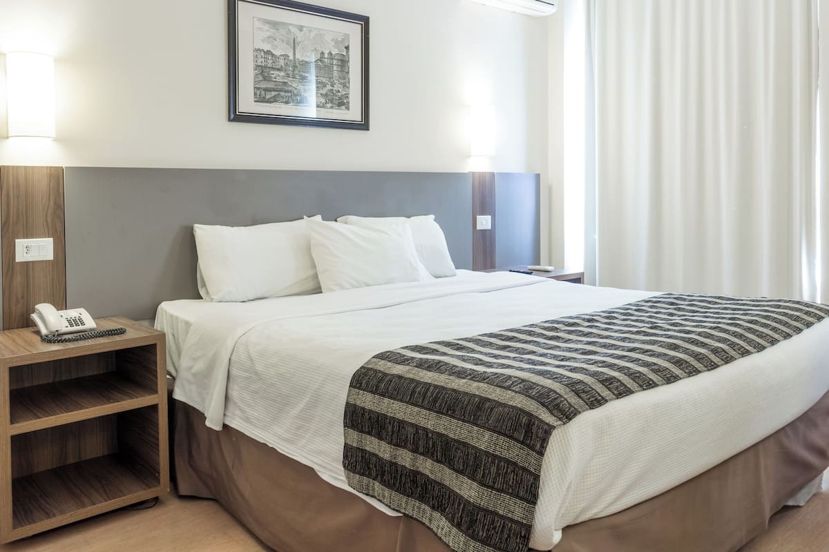 Una cama bien hecha con sábanas blancas, cabecera marrón y manta a rayas. Hay una mesita de noche con una lámpara y un teléfono. Encima de la cama, hay un cuadro enmarcado en la pared.