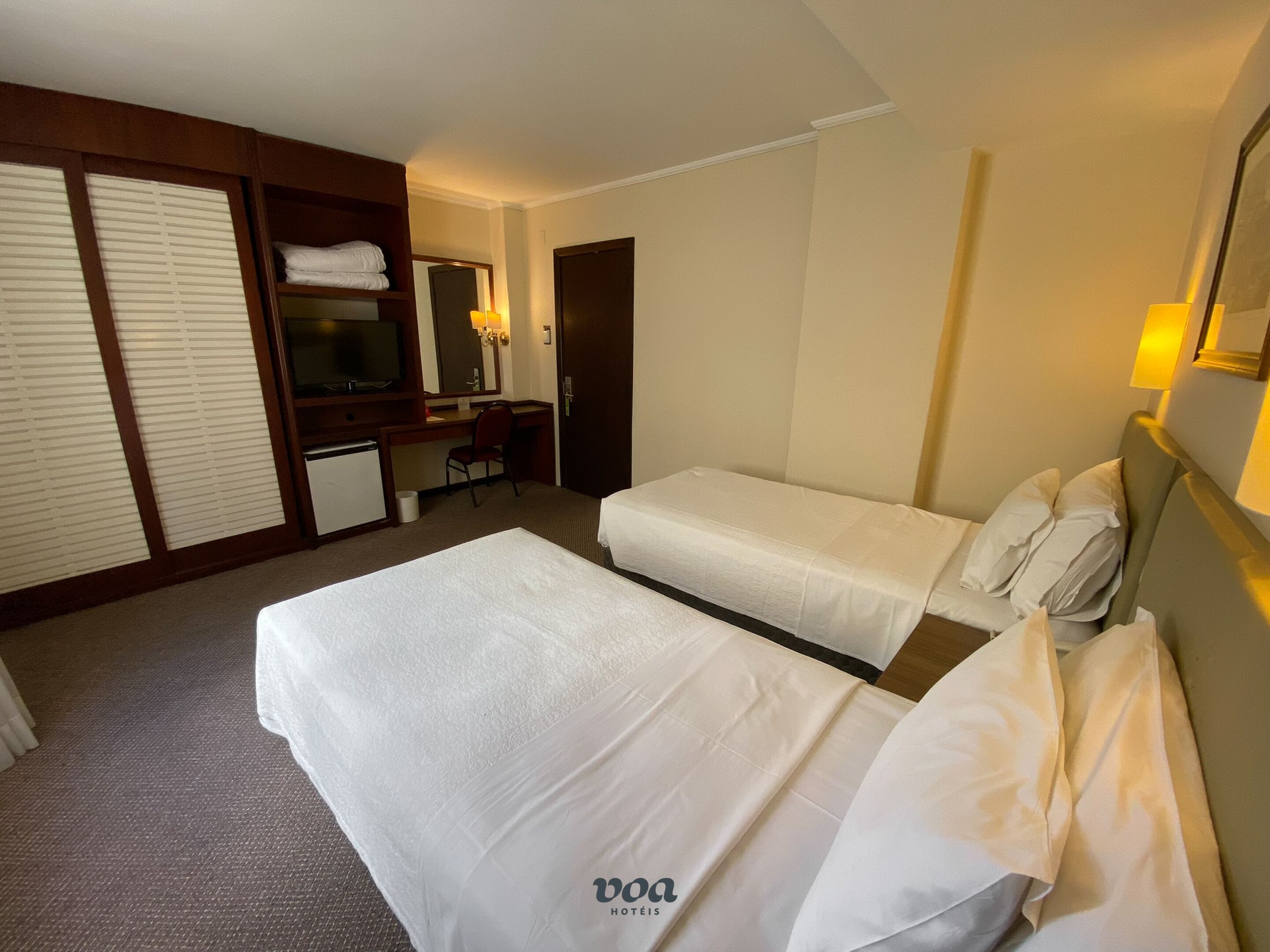 A hotel room with two beds, a desk, a chair, a TV, and a wardrobe.