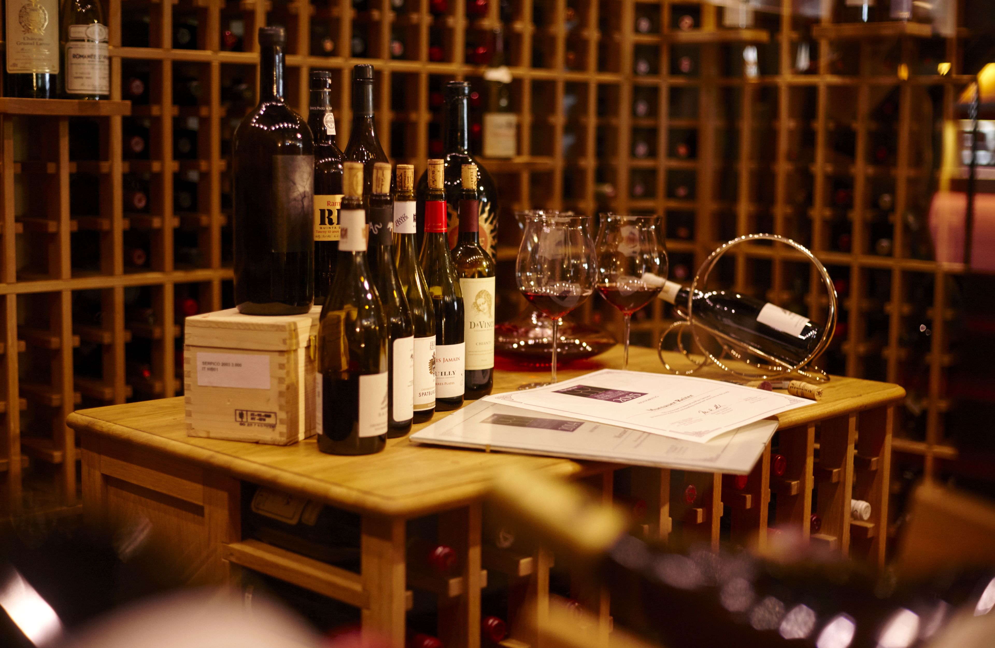 A wooden table with wine bottles and glasses, surrounded by a wine rack in the background.