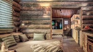 Interior de cabana de madeira com uma cama aconchegante, paredes de madeira, lareira de pedra e uma área de cozinha rústica.