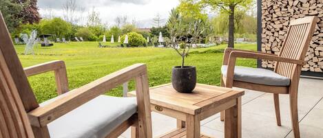 A wooden outdoor seating area with two chairs and a table, a potted plant, and a backdrop of neatly stacked firewood.
