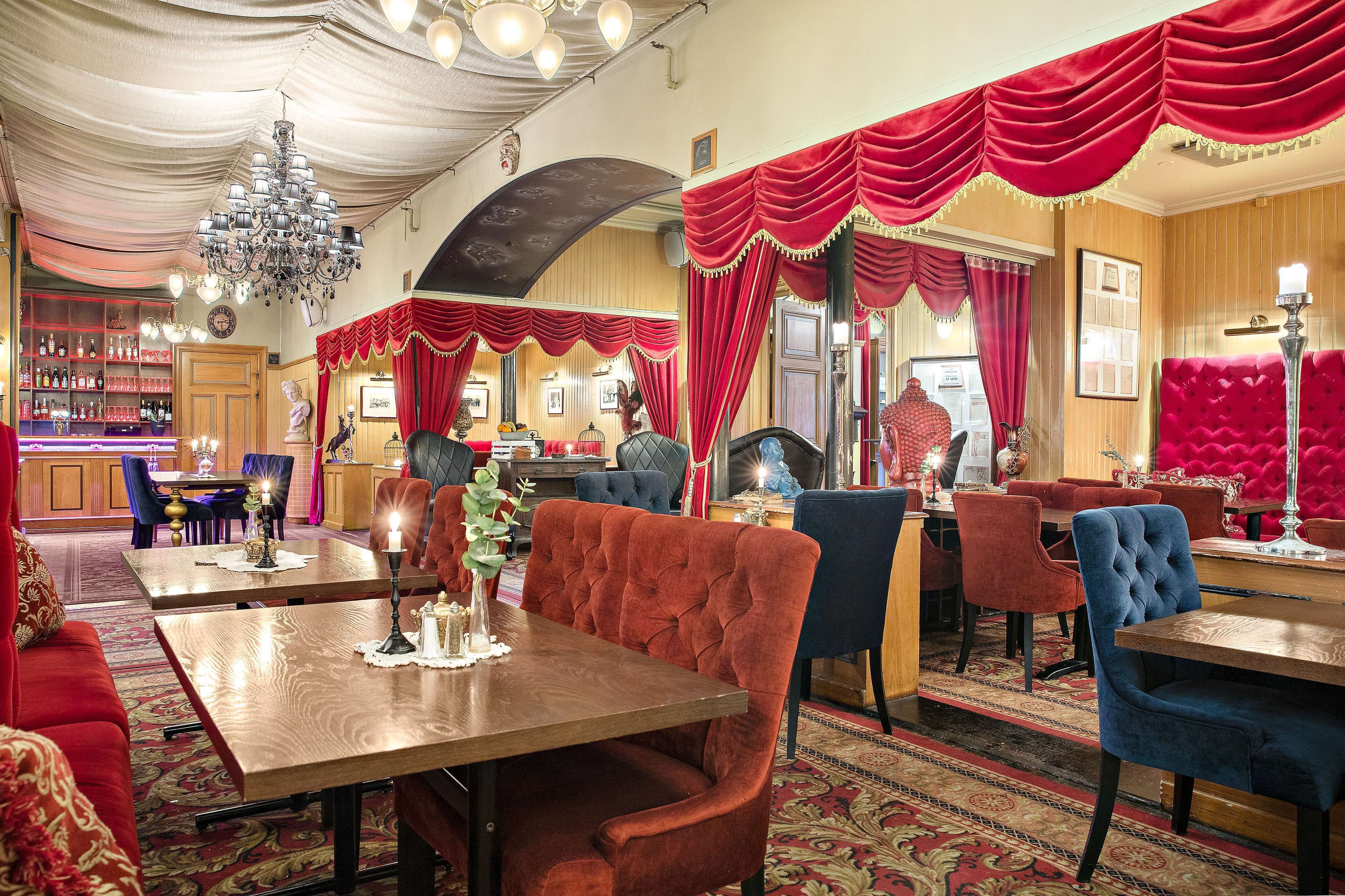 Un restaurante con sillas tapizadas en rojo y azul, un candelabro y una zona de bar al fondo.