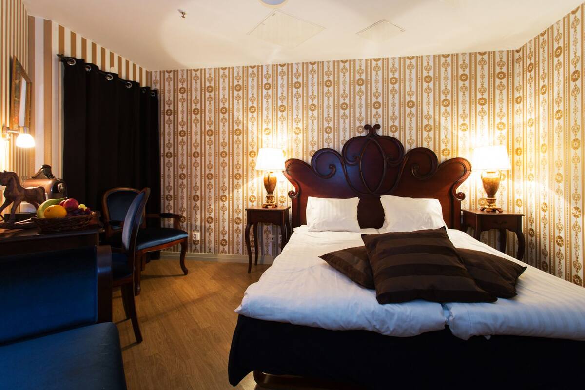 Ein Hotelzimmer mit einem großen Bett, zwei Nachttischen mit Lampen, einem Sessel, einem Schreibtisch mit Obst und einer Pferdestatue, sowie rautenmusterter Tapete.