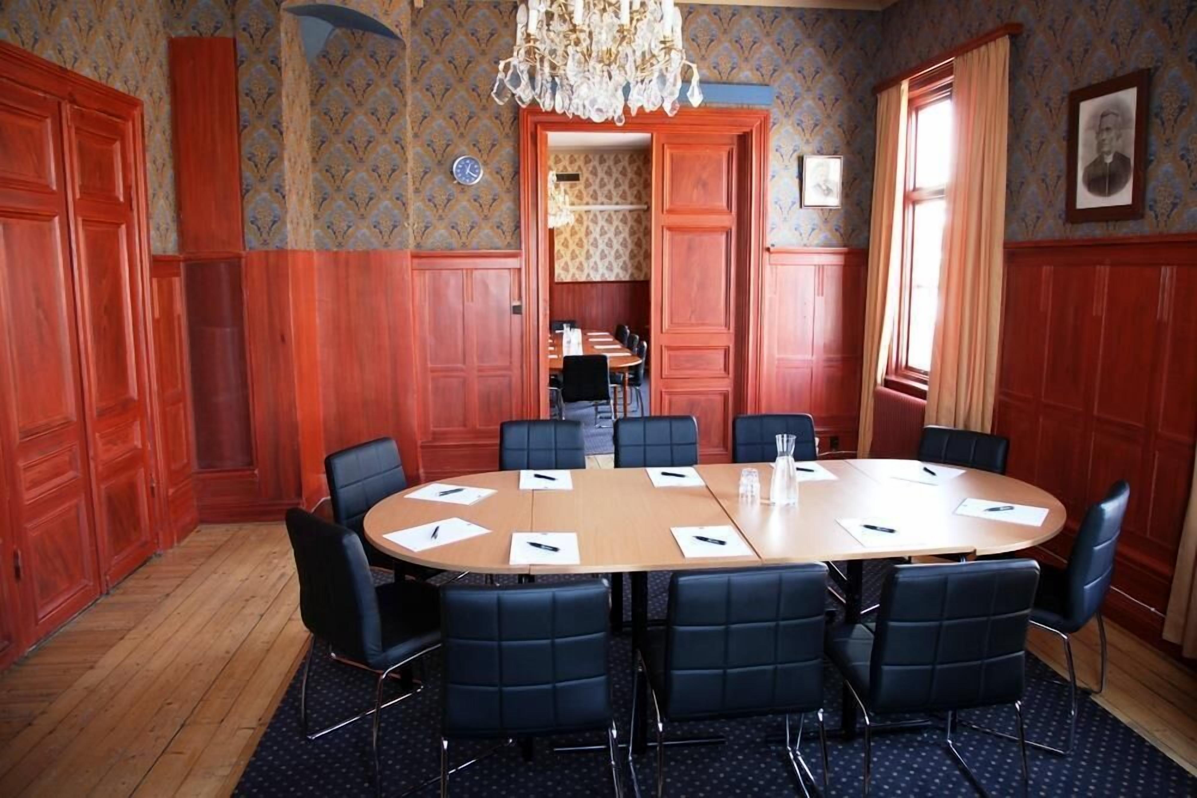 Una sala de conferencias con una mesa redonda, sillas negras, paneles de madera y un candelabro.